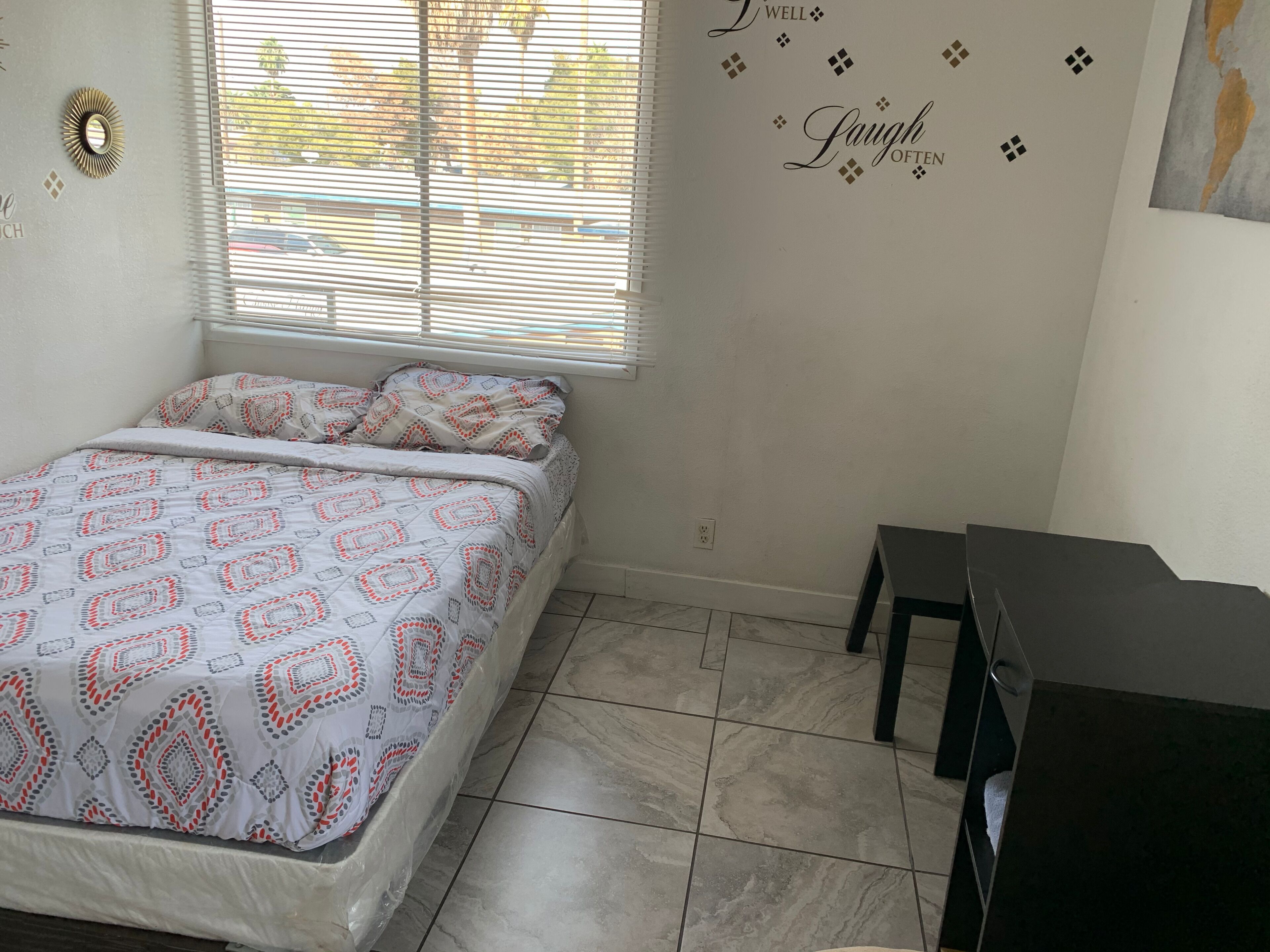 Une chambre à coucher avec un lit, une fenêtre avec des stores, un élément décoratif mural et une petite table.