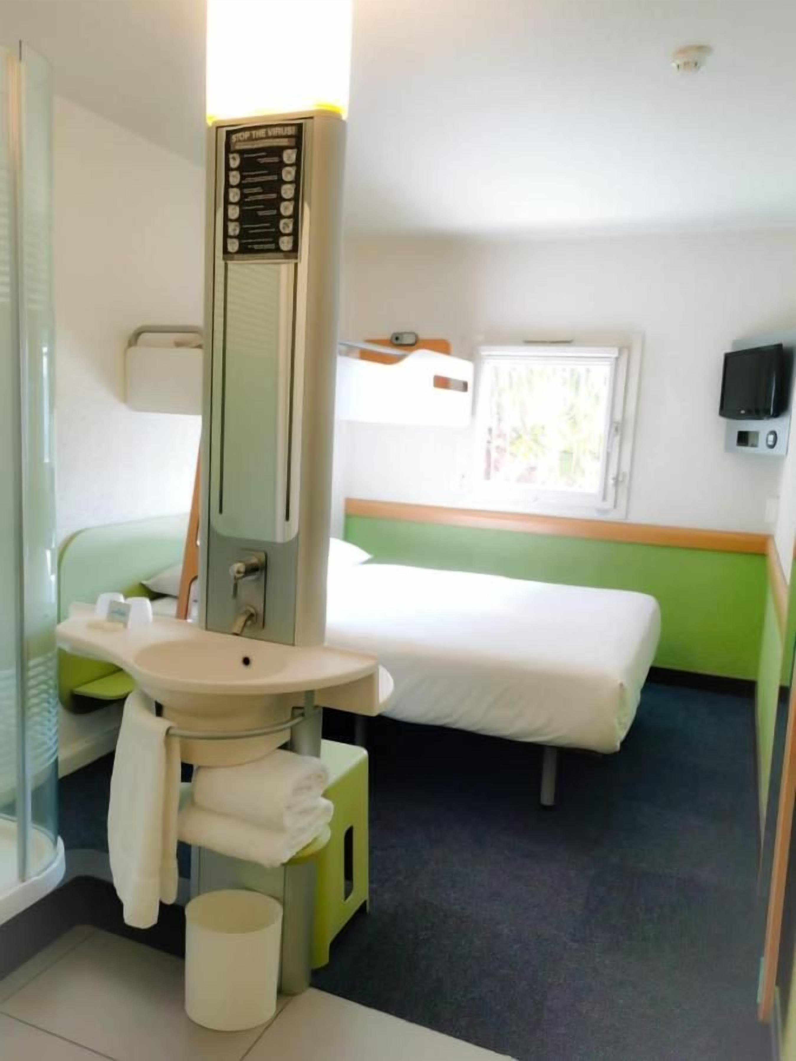 A hotel room with a bed, a bedside table, a sink, and a mirror.