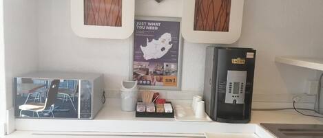Cozinha equipada com máquina de café, micro-ondas e frigorífico. Duas imagens emolduradas na parede e um cartaz com texto.