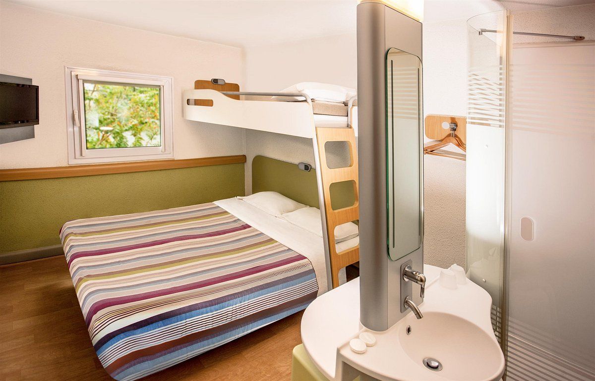 A bunk bed with a striped bedspread, a bedside table with a mirror, a sink, and a window with a view of greenery.