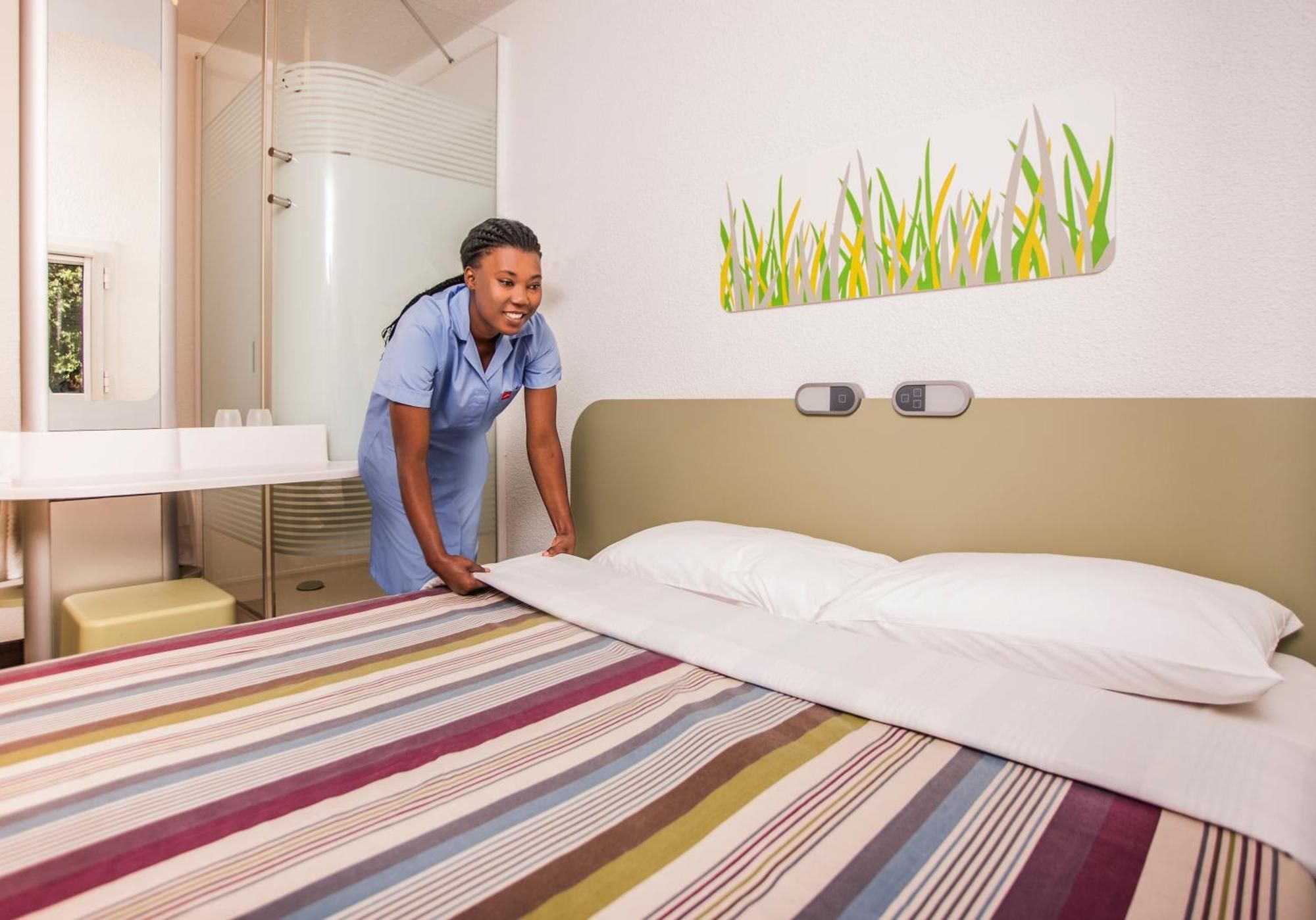 Habitación de hotel con cama, mesita de noche, espejo y panel de control en la pared.