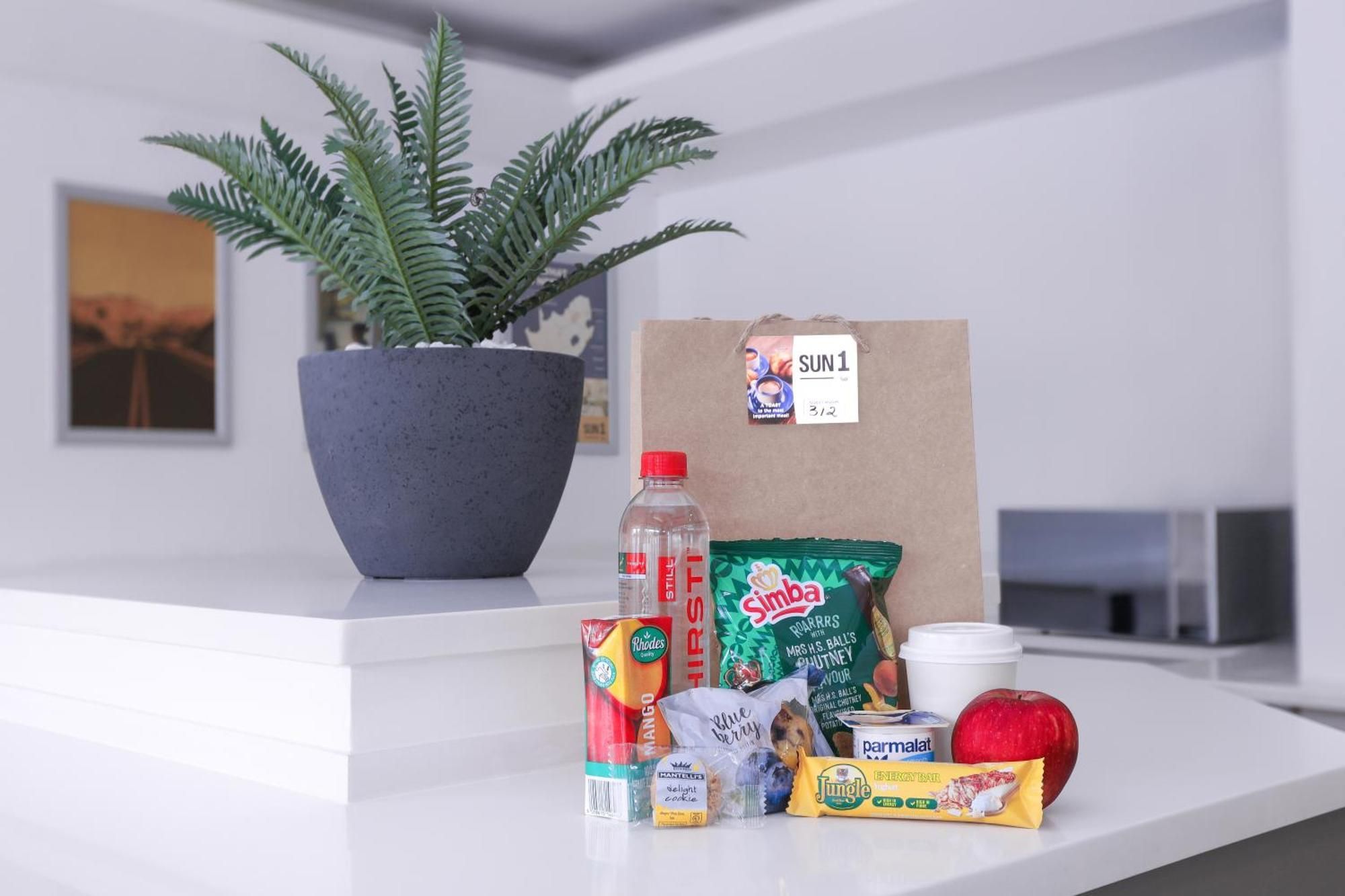 A kitchen counter with a potted plant, a brown paper bag, and various food items including a bottle of water, a can of mango, a packet of chips, a jar of peanut butter, a packet of instant oatmeal, a red apple, and a container of yogurt.