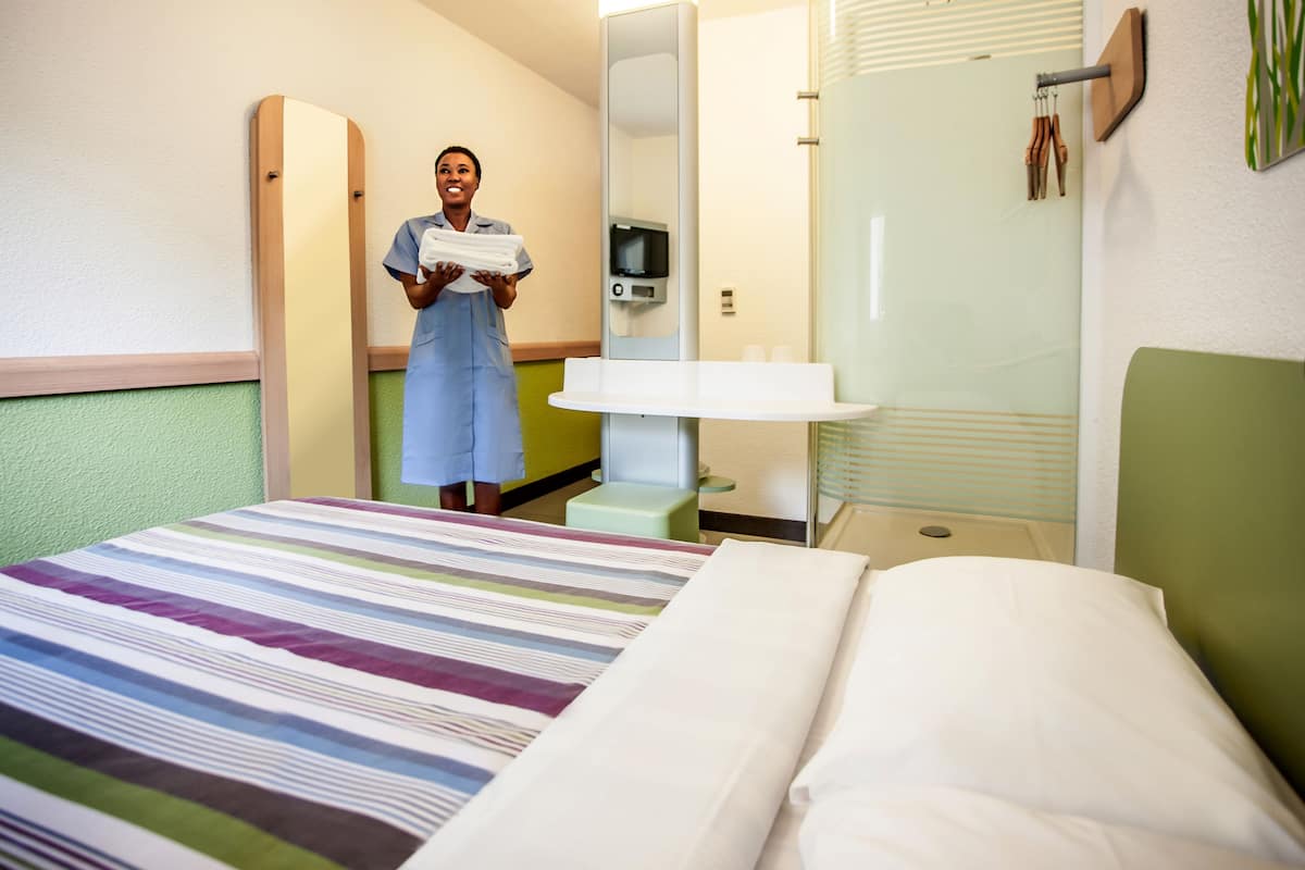 A hotel room with a bed, a bedside table, a mirror, and a bathroom with a sink and towel rack.