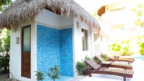 Un petit bâtiment avec un toit de chaume et un mur décoré de mosaïques bleues. On y trouve des chaises longues en bois près d’une piscine.