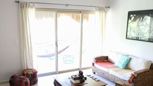 A room with a wicker sofa, a small table, a colorful quilted chair, and a large window with a view of a hammock and a thatched roof structure.