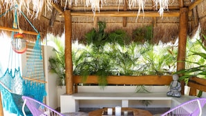 A thatched-roof outdoor seating area with a bench, a hanging hammock, and a statue.