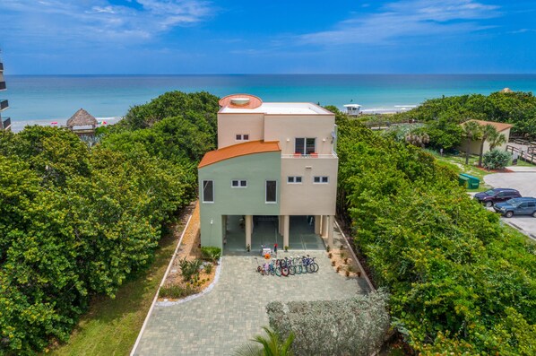 Un edificio moderno con techo curvo, rodeado de abundante vegetación, cerca de una playa con aguas azul cristalino.