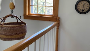 A wooden staircase with a white railing, a hanging basket, a window with a view of trees, and a wall-mounted clock. - Charming Post and Beam Cottage! Walk to the beach, Tennis, Bike Path, & Shuttle (Nantucket)
