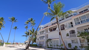 A beachfront hotel with multiple balconies, palm trees, and a clear blue sky. - Beachfront 2BR Apt · Los Corales, Bávaro · Utilities Included (Punta Cana)