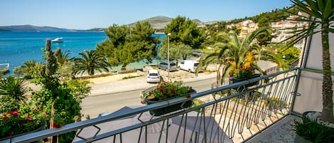 A balcony with a view of the sea, parked cars, and greenery.