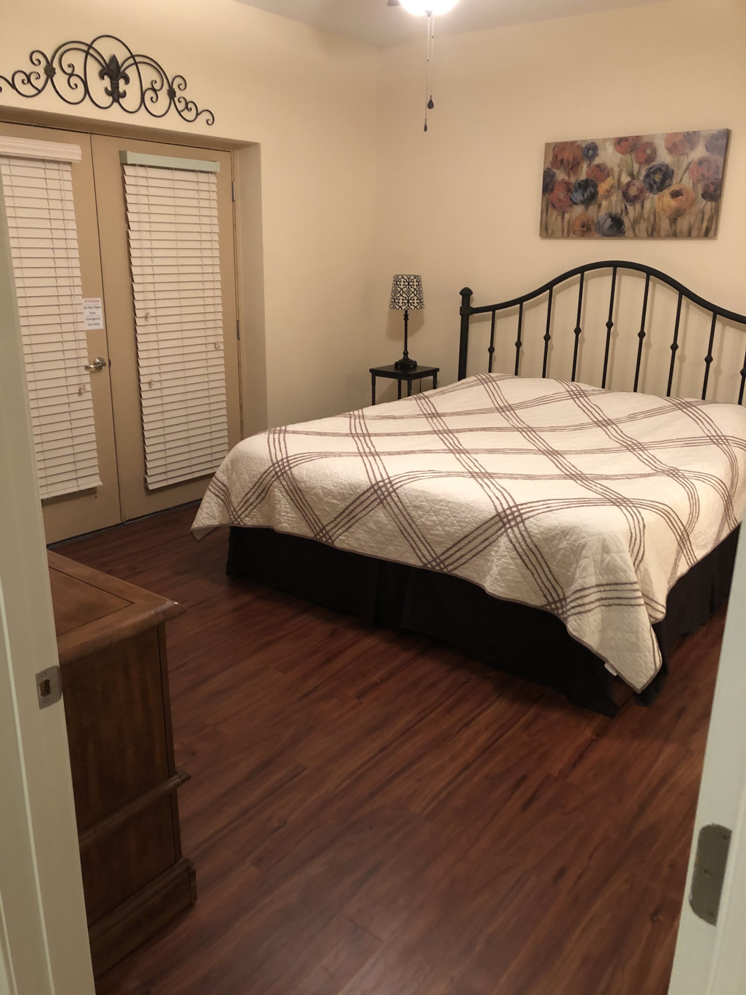 A bedroom with a bed, wooden floor, a nightstand, a wall-mounted lamp, and a painting.