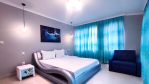 Ein modernes Schlafzimmer mit einem gebogenen Bett, einem kleinen Nachttisch, einem Sessel und einem großen Fenster mit blauen Vorhängen.