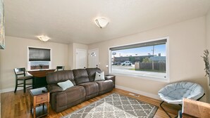 A living room with a brown leather sofa, a dining table with chairs, a large window with a view of the street, and a gray patterned rug. - This is Boise's Gem! (Boise)