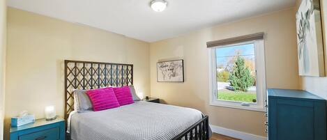 Un dormitorio con cama, mesita de noche, cómoda, ventana con vistas y una lámpara en el techo.