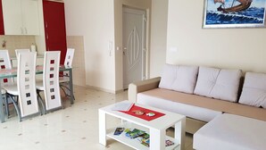 Un salon moderne avec un canapé beige, une table basse blanche sur laquelle sont posés des magazines, et un coin repas avec des chaises blanches et un buffet rouge.