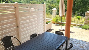 Une terrasse avec une table et des chaises, une clôture blanche et un jardin agrémenté de plantes et d’une structure en pierre.