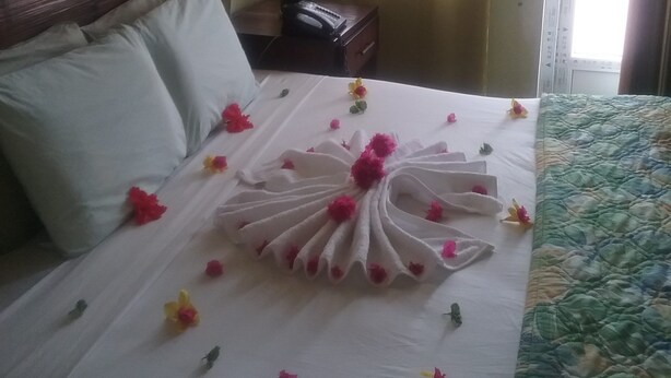 A neatly made bed with a floral arrangement, a bedside lamp, and a window with curtains. - Aanola Villas 6B Quiet boulevard (Charlotte)