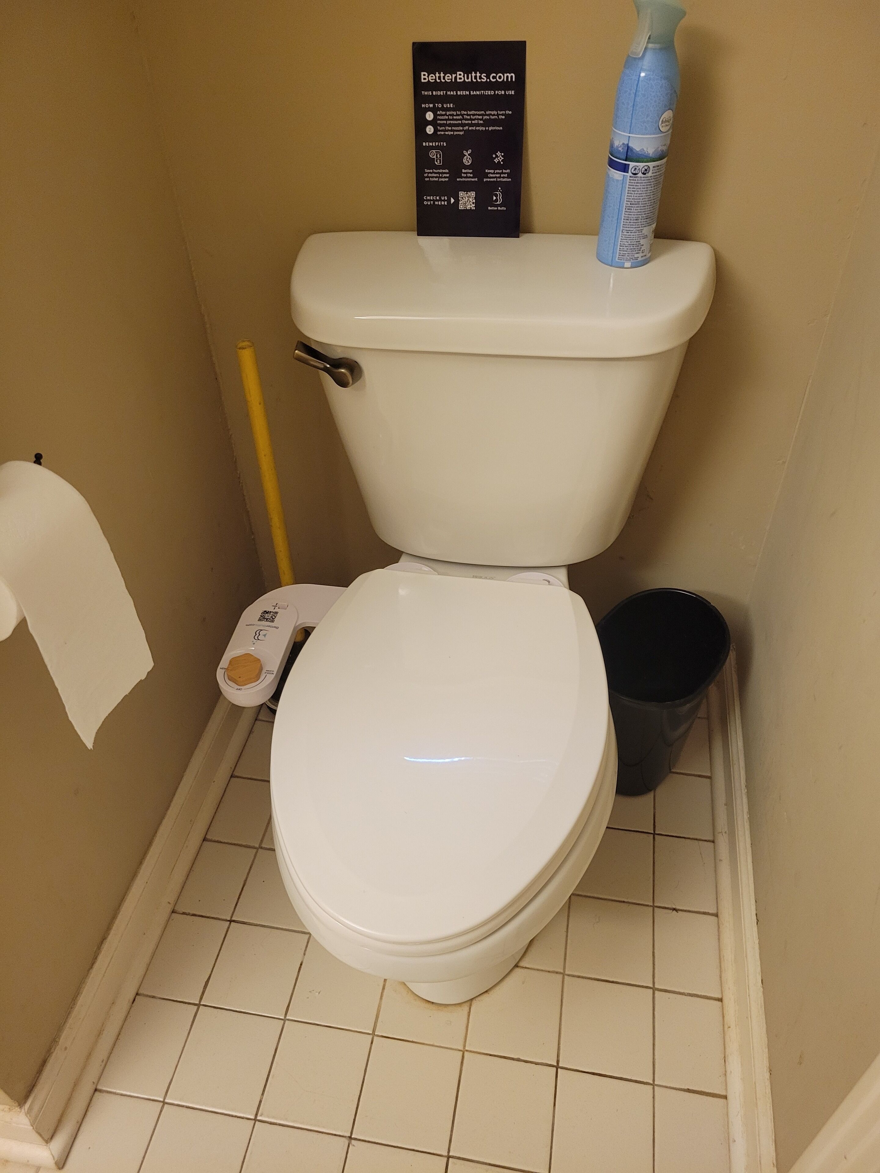 Um vaso sanitário branco com uma placa em cima, um rolo de papel higiênico, um borrifador de produto de limpeza e uma lixeira.