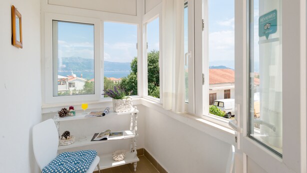 Une pièce aux murs blancs, une chaise blanche ornée d’un coussin à pois, une petite étagère blanche décorée de divers objets, et de grandes fenêtres offrant une vue sur les bâtiments et les montagnes.