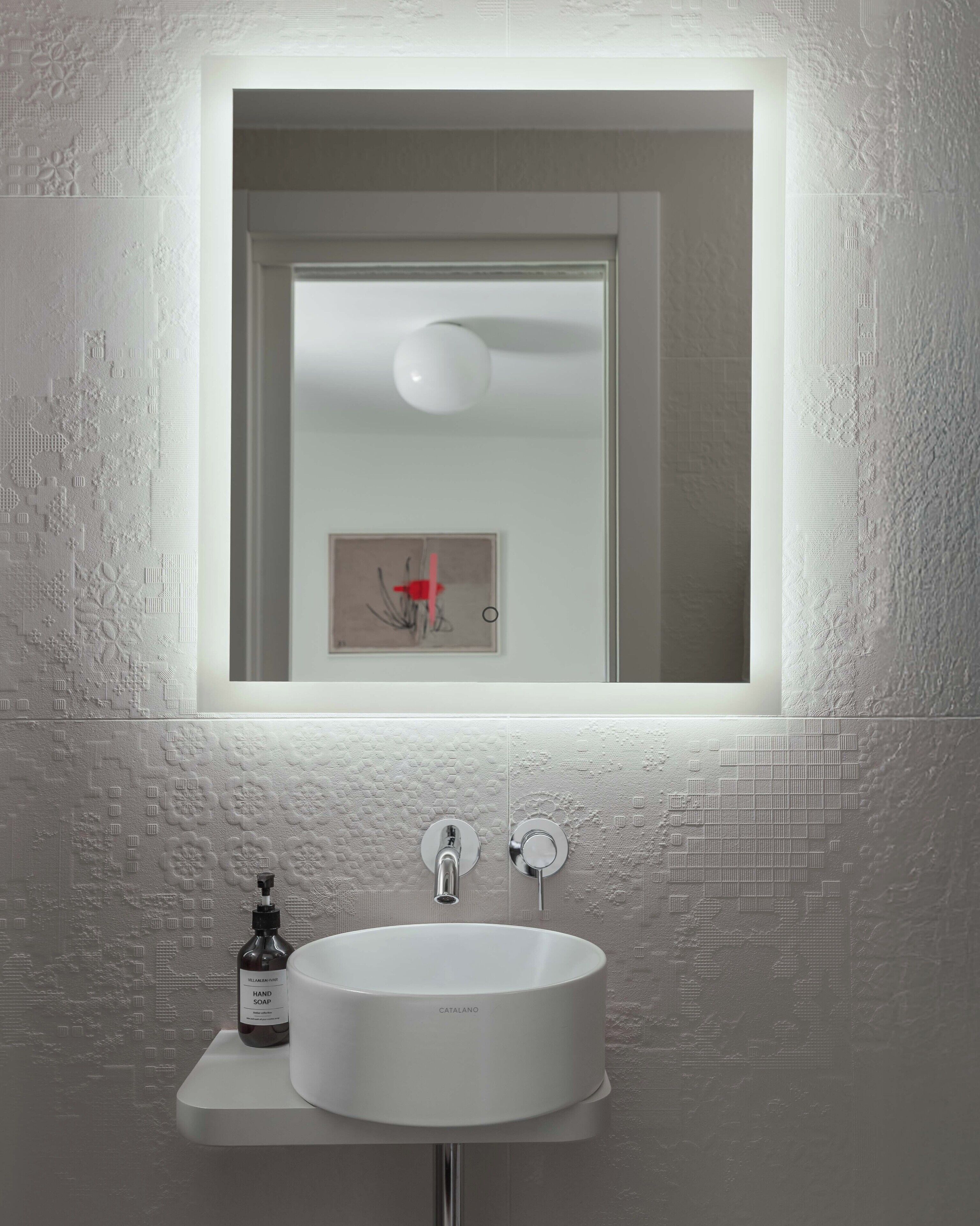 A bathroom with a white sink, a hand soap dispenser, and a mirror with a framed artwork.