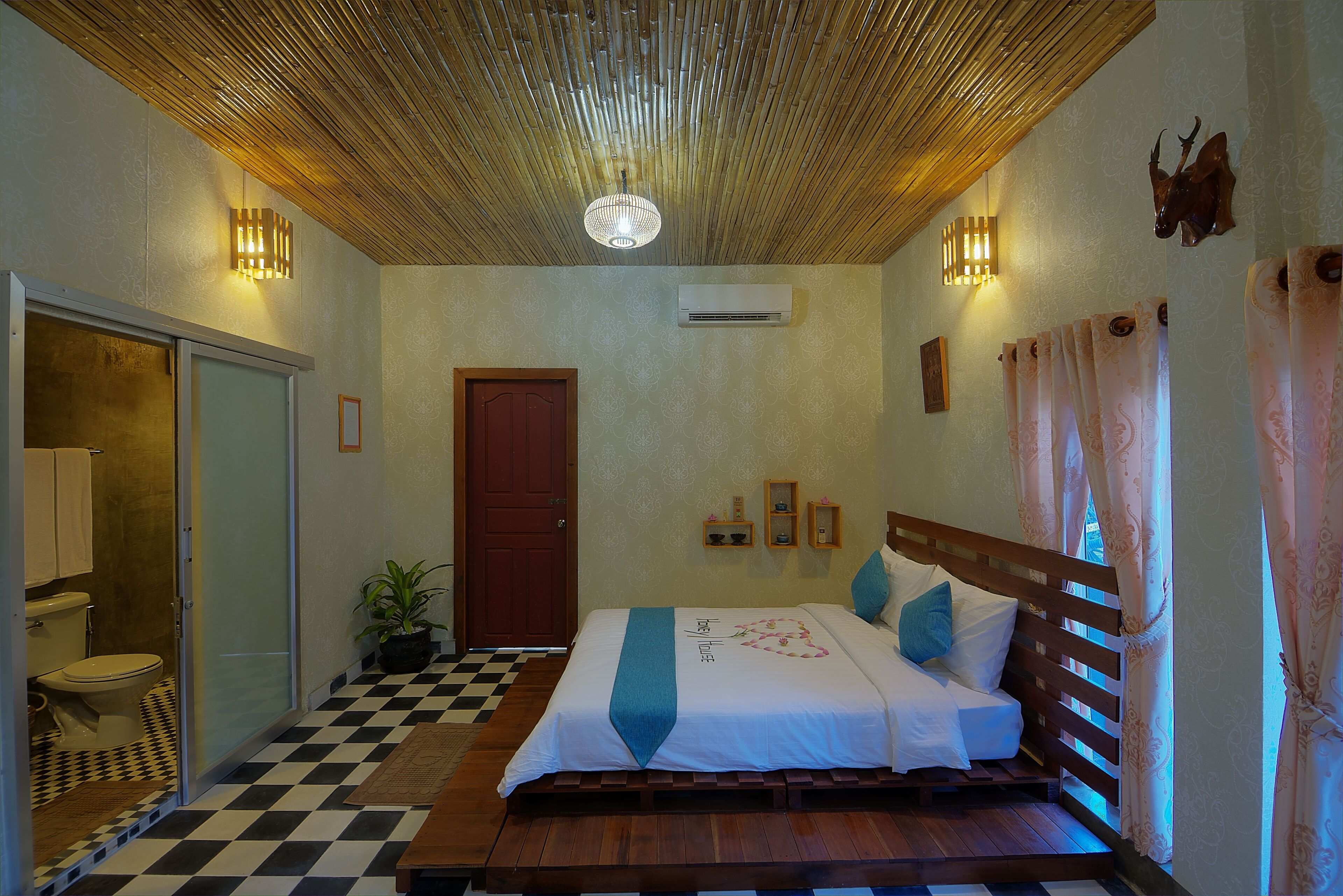 Hotelkamer met een zwart-wit betegelde vloer, een bed met een houten hoofdbord, een kamerplant, een wandairconditioner en een deur.