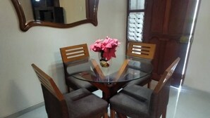 Un comedor con una mesa redonda de vidrio, cuatro sillas de madera, un jarrón con flores rosas, un espejo grande y una puerta de madera con paneles de vidrio.