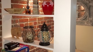 Un salon chaleureux avec un mur en briques, des étagères ornées d’objets décoratifs, un canapé et un tapis à motifs.