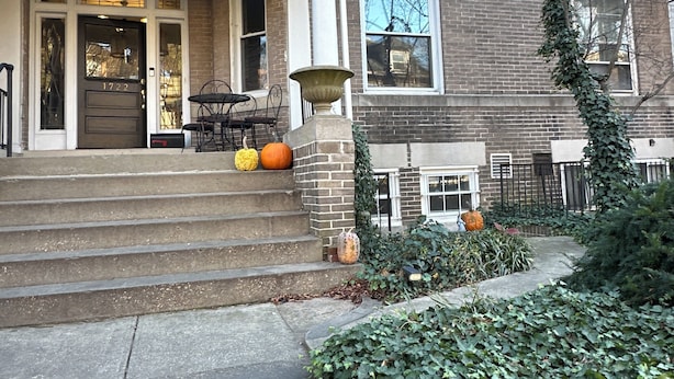 Un bâtiment en briques de deux étages, doté d’un porche à l’avant, d’un escalier menant à l’entrée et d’un petit jardin où l’on peut voir des citrouilles et du lierre.