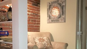 Un salon avec un mur en briques, un fauteuil beige agrémenté d’un coussin à motifs floraux et un pouf assorti. On y trouve une bibliothèque avec des objets décoratifs et un miroir encadré accroché au mur.