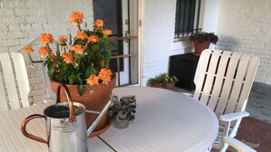 Une table blanche pour l’extérieur, avec un arrosoir en métal et une plante en pot aux fleurs orange.
