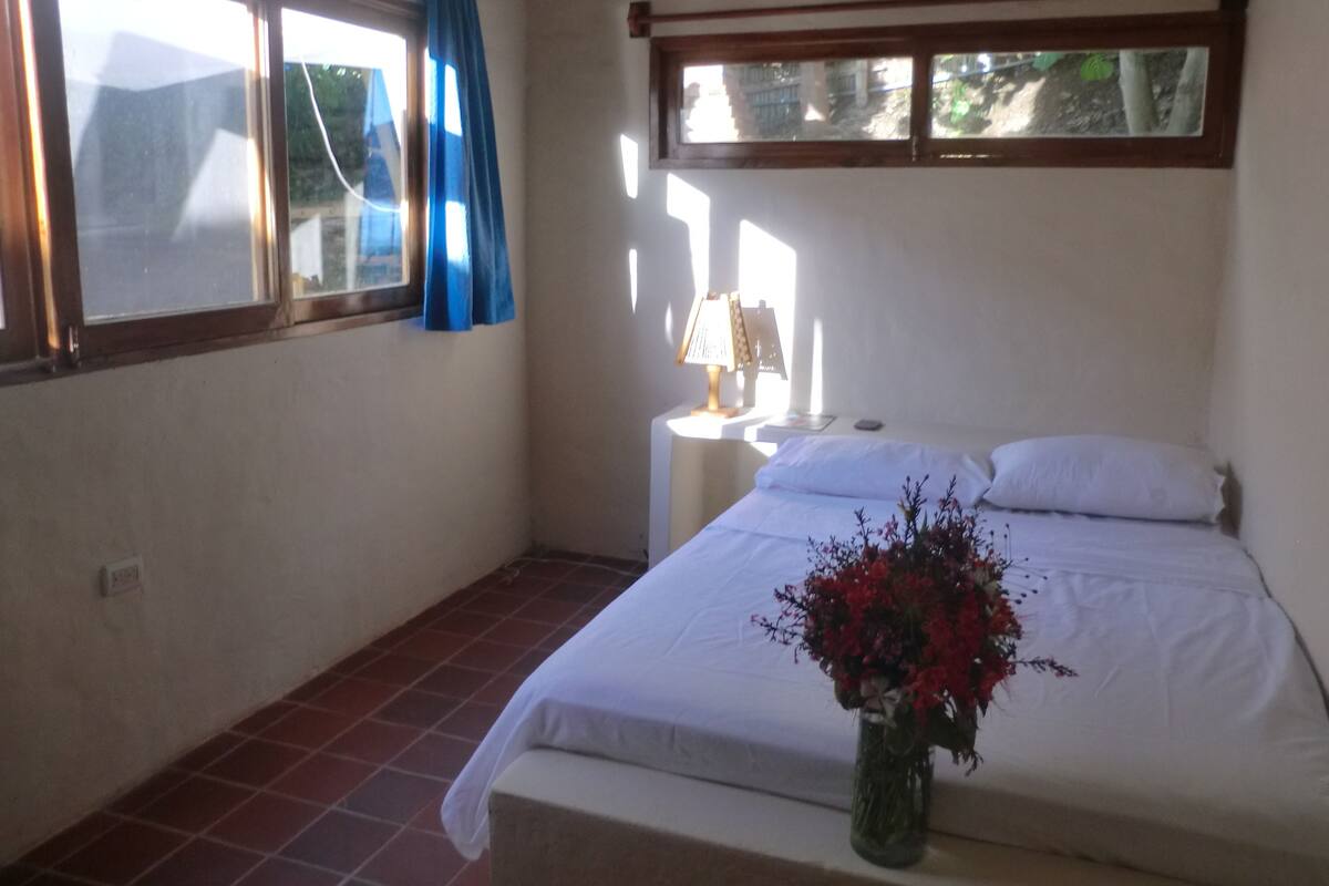 A bedroom with a bed, a nightstand, a lamp, a window with curtains, and a vase with flowers.