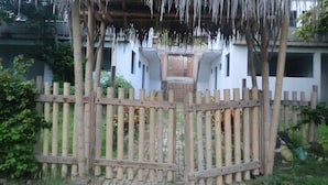 Un mirador de madera con techo de paja, rodeado por una valla de bambú, y un edificio blanco con entrada central al fondo.