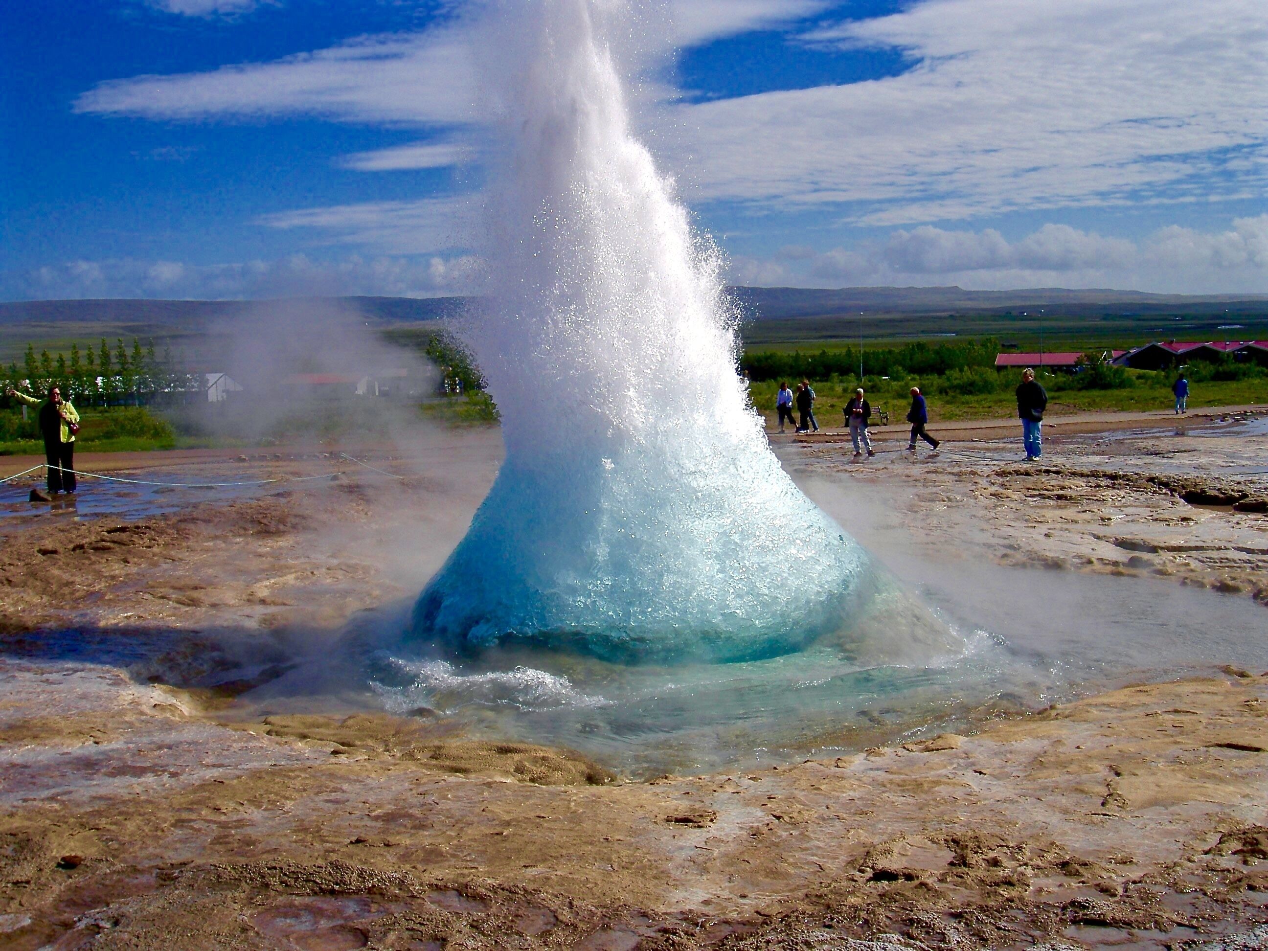A geyser with a clear water stream, surrounded by a muddy terrain and a few people observing.
