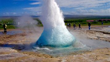 A geyser with a clear water stream, surrounded by a muddy terrain and a few people observing.