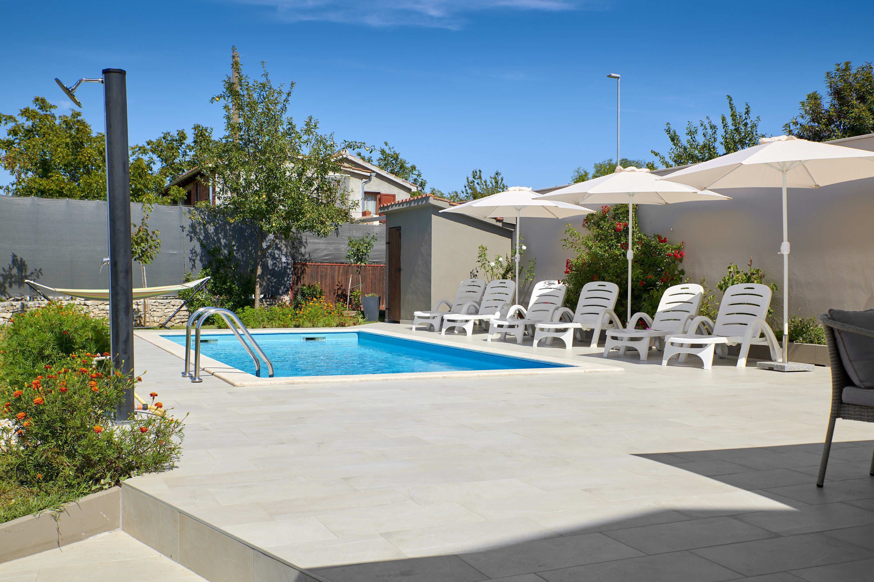 Ein Swimmingpool mit weißen Liegestühlen und Sonnenschirmen, umgeben von viel Grün und einem klaren Himmel.