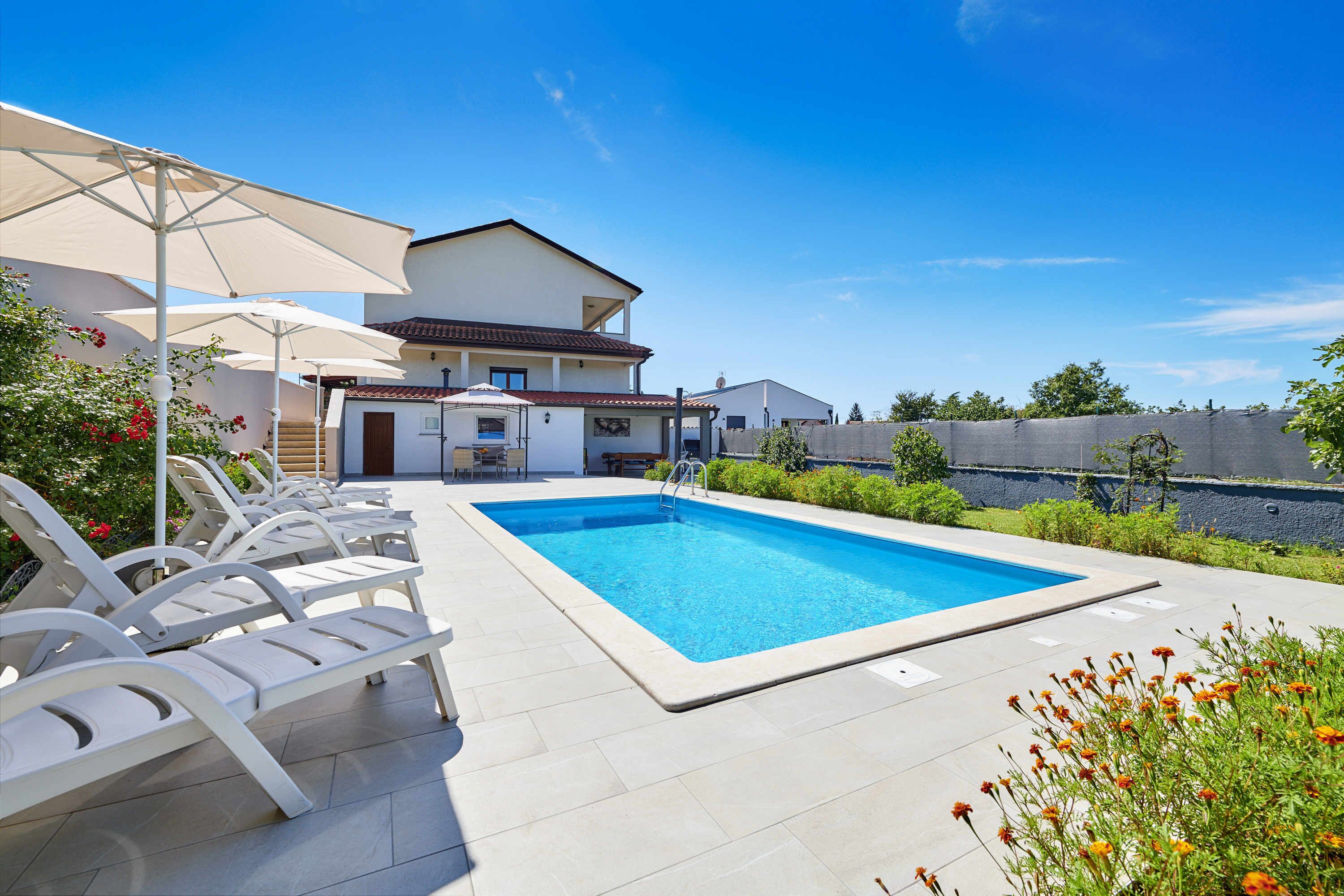 A swimming pool with lounge chairs and umbrellas, surrounded by a house and greenery.