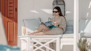 A person sitting on a white outdoor bench reading a book, with a drink on the table and a wooden door in the background.