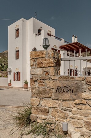 A stone wall with a sign reading 'Villa of Roses' in front of a white building with arched windows and a red roof.