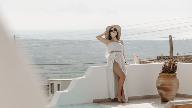 A woman in a white dress and hat sitting by a pool with a view of the landscape.