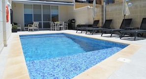 Una piscina rettangolare con piastrelle blu a mosaico, circondata da piastrelle color beige e confortevoli poltrone da relax nere.