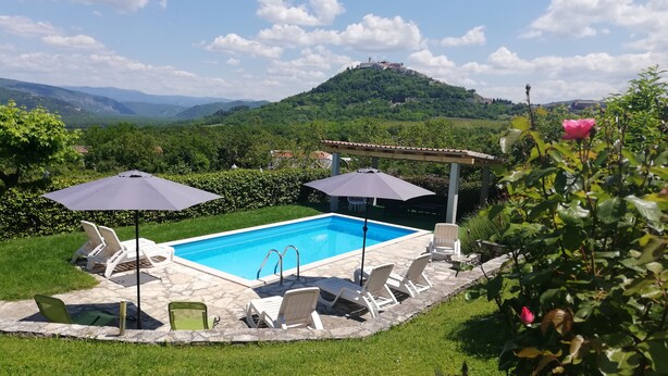 A swimming pool with lounge chairs and umbrellas, surrounded by greenery and a view of hills.