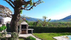 A stone outdoor kitchen with a chimney, surrounded by a well-maintained lawn and a large tree.