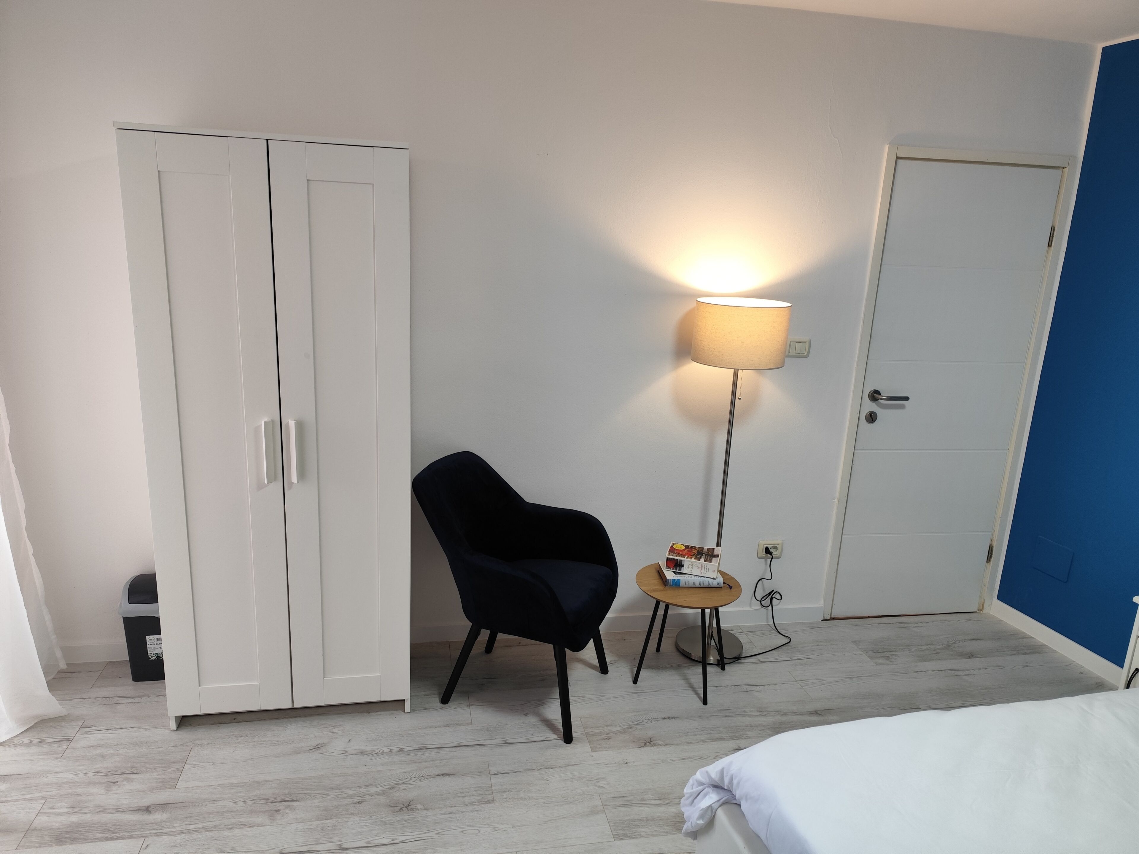A bedroom with a bed, a wardrobe, a chair, a floor lamp, and a small table with a book.