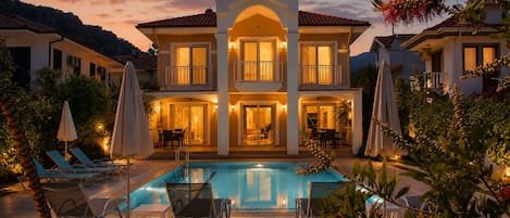 A two-story house with a swimming pool, outdoor seating, and a well-lit entrance.