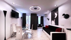 A modern living room with a sofa, a dining table with chairs, a TV mounted on the wall, and a large window with curtains.