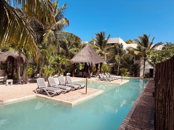 A pool area with lounge chairs and a thatched hut, surrounded by palm trees and a clear sky. - Apartment in Playa Chaca Complex near downtown Progreso  (Progreso)