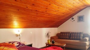 Ein gemütliches Zimmer mit Holzbalkendecke, einem Bett mit violett-orange gestreifter Decke, einer roten Tagesdecke, einem kleinen Tisch mit Pflanze und einem Sofa mit gemusterten Kissen.