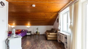 Ein gemütliches Schlafzimmer im Dachgeschoss mit Holzbalkendecke, einem Bett mit bunter Decke, einem kleinen Tisch mit Pflanze, einer Couch und einem Fenster mit Vorhängen.