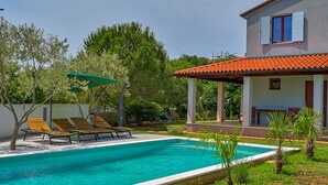 Ein Schwimmbecken mit Liegestühlen, eine überdachte Terrasse und ein Gebäude mit roten Ziegeldach.