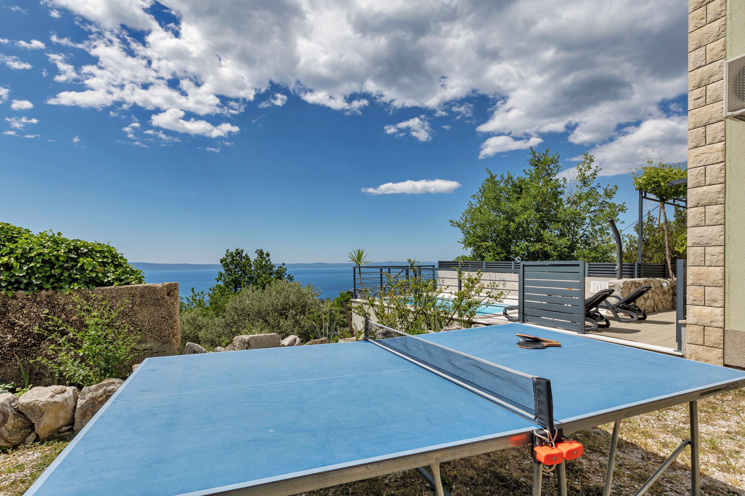 A ping pong table with a net, a stone wall, and a view of the sea.